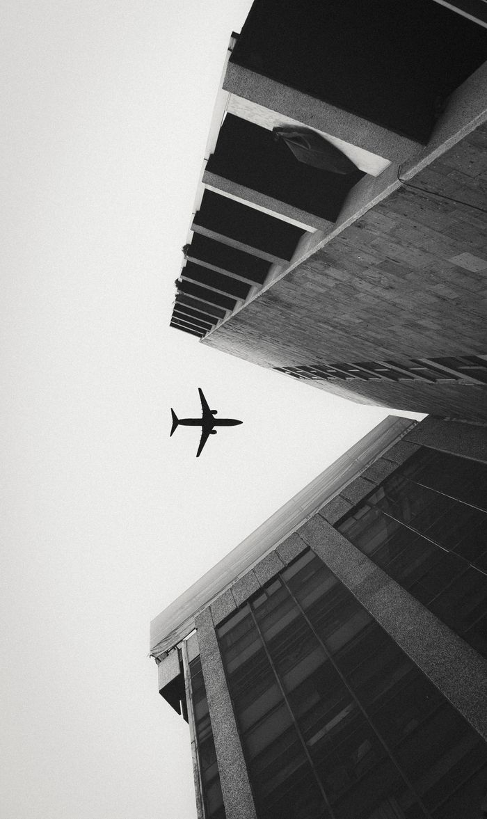 About Monochrome photo of an airplane flying above modern city buildings.