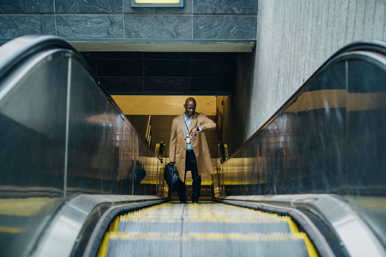 About High angle full length pensive African American businessman in formal clothes with laptop case riding escalator and checking time on wristwatch while leaving metro station