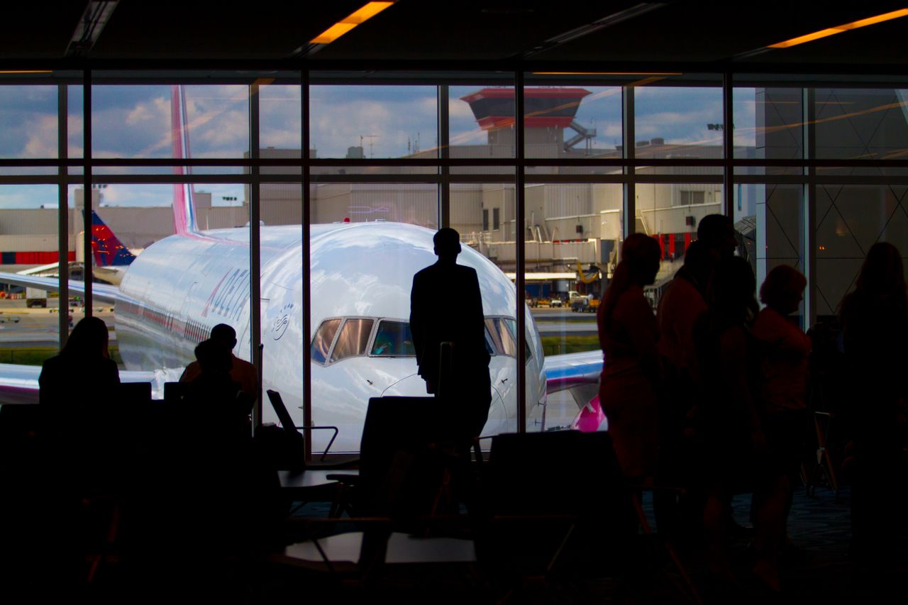 Home Silhouettes of travelers waiting at an airport terminal with an airplane visible through the window.
