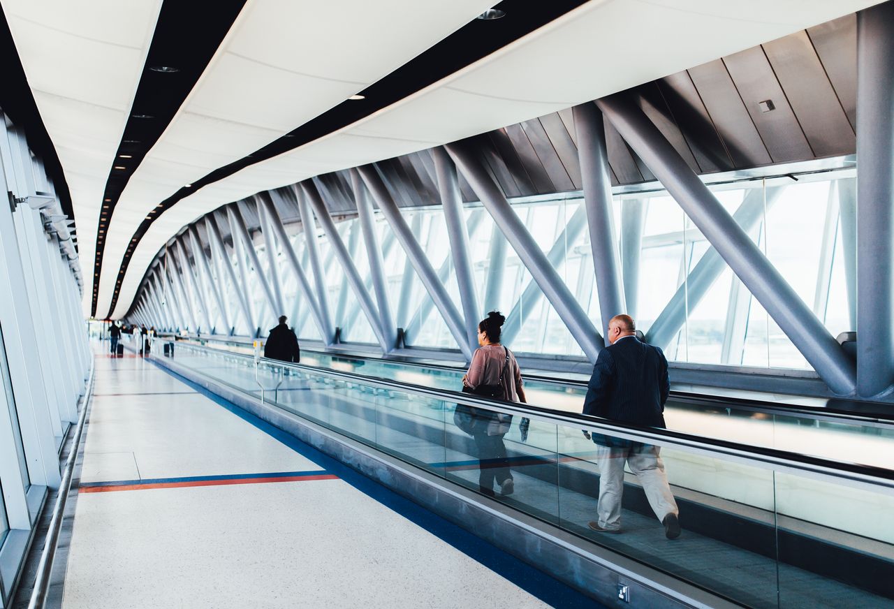 Home Travelers walking through a bright modern airport corridor with glass walls.